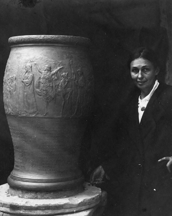 Black and white photo of woman facing the camera next to large urn on platform beside her