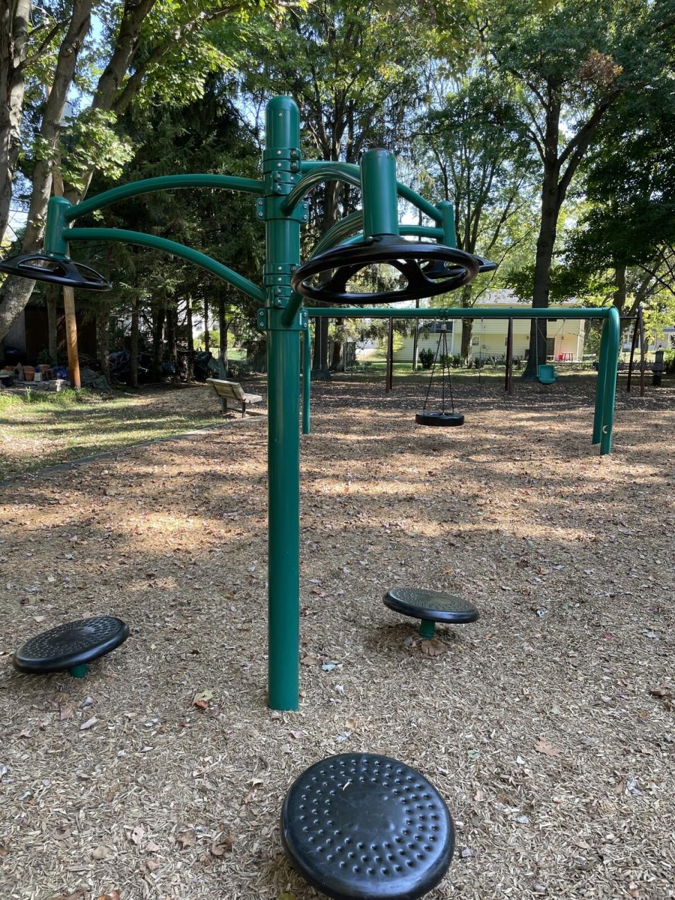 Color photo of green fitness station and swings in background on mulched playground