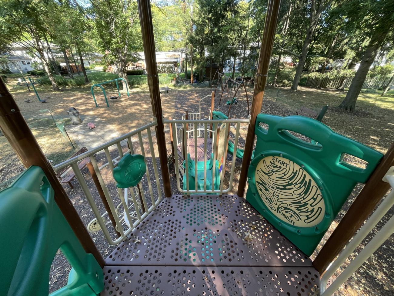 Color photograph looking down over railing at top of climbing gym and slide with mulched park and trees in background