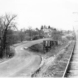 Black and white photo of a dirt road winding through leafless trees, with a railway track visible at the right and an unidentified platform or wooden structure in between the road and railway