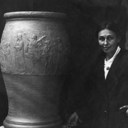 Black and white photo of woman facing the camera next to large urn on platform beside her
