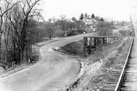 Black and white photo of a dirt road winding through leafless trees, with a railway track visible at the right and an unidentified platform or wooden structure in between the road and railway
