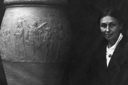 Black and white photo of woman facing the camera next to large urn on platform beside her