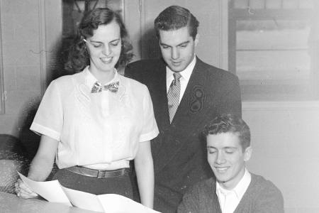 Black and white photo of three people looking at piano