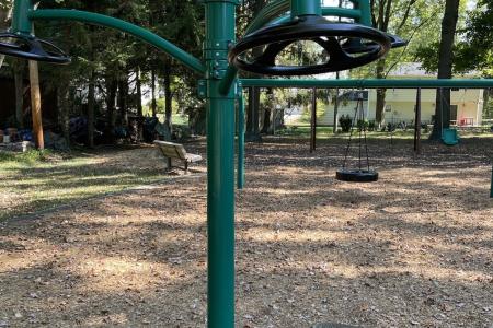 Color photo of green fitness station and swings in background on mulched playground