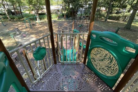 Color photograph looking down over railing at top of climbing gym and slide with mulched park and trees in background